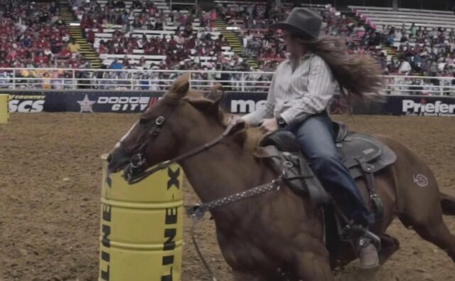 A barrel racer in a rodeo arena makes a tight turn around a yellow barrel on a brown horse, displaying focused determination.